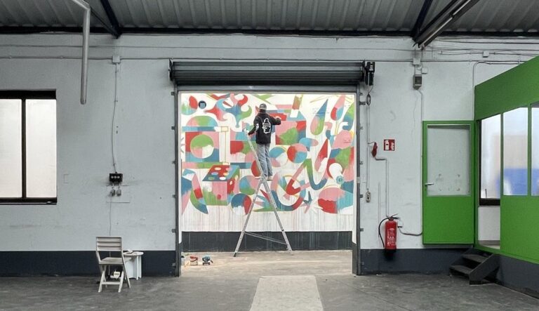 Ein Mann steht in einer Industriehalle auf einer Leiter. Er malt ein Graffitti an eine Wand.