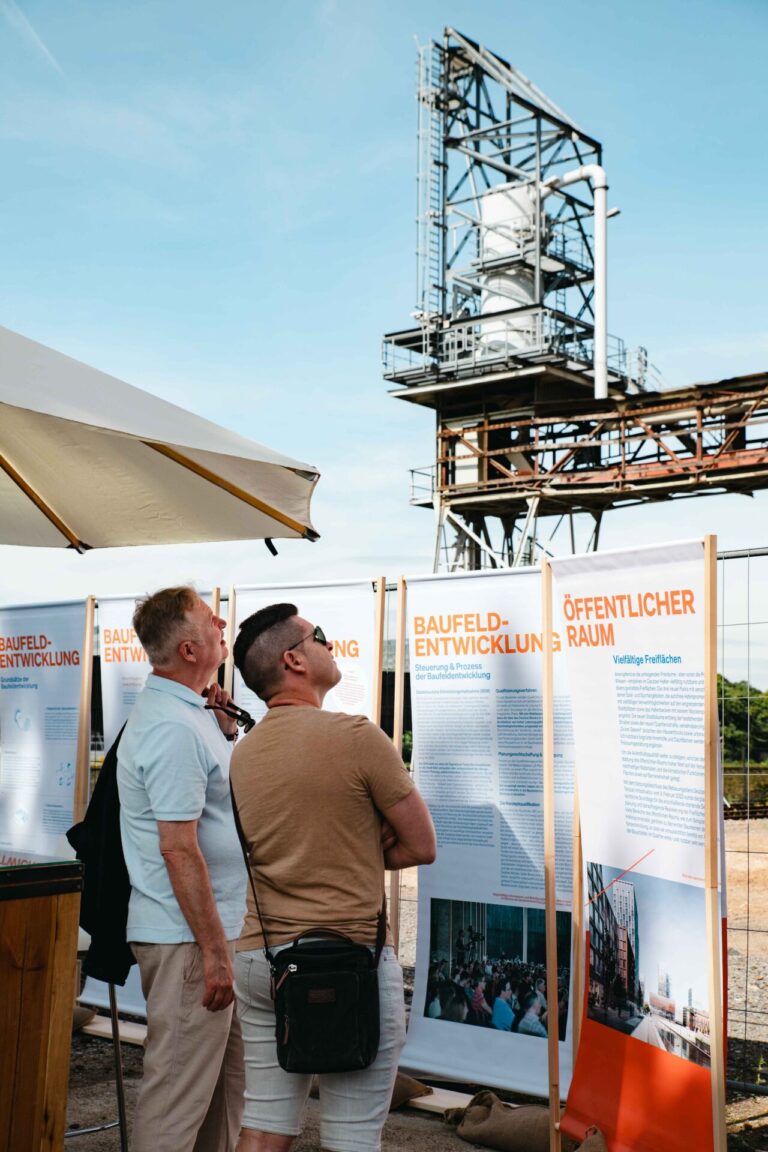 Zwei Besucher betrachten beim Hafentag 2023 Informationstafeln zur Entwicklung des Deutzer Hafens. Im Hintergrund erhebt sich eine historische Industrieanlage.
