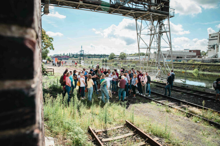 Besucherinnen und Besucher stehen bei einer Führung im Deutzer Hafen unter einer historischen Industrieanlage zusammen. Daneben befinden sich alte Gleise und das Hafenbecken.