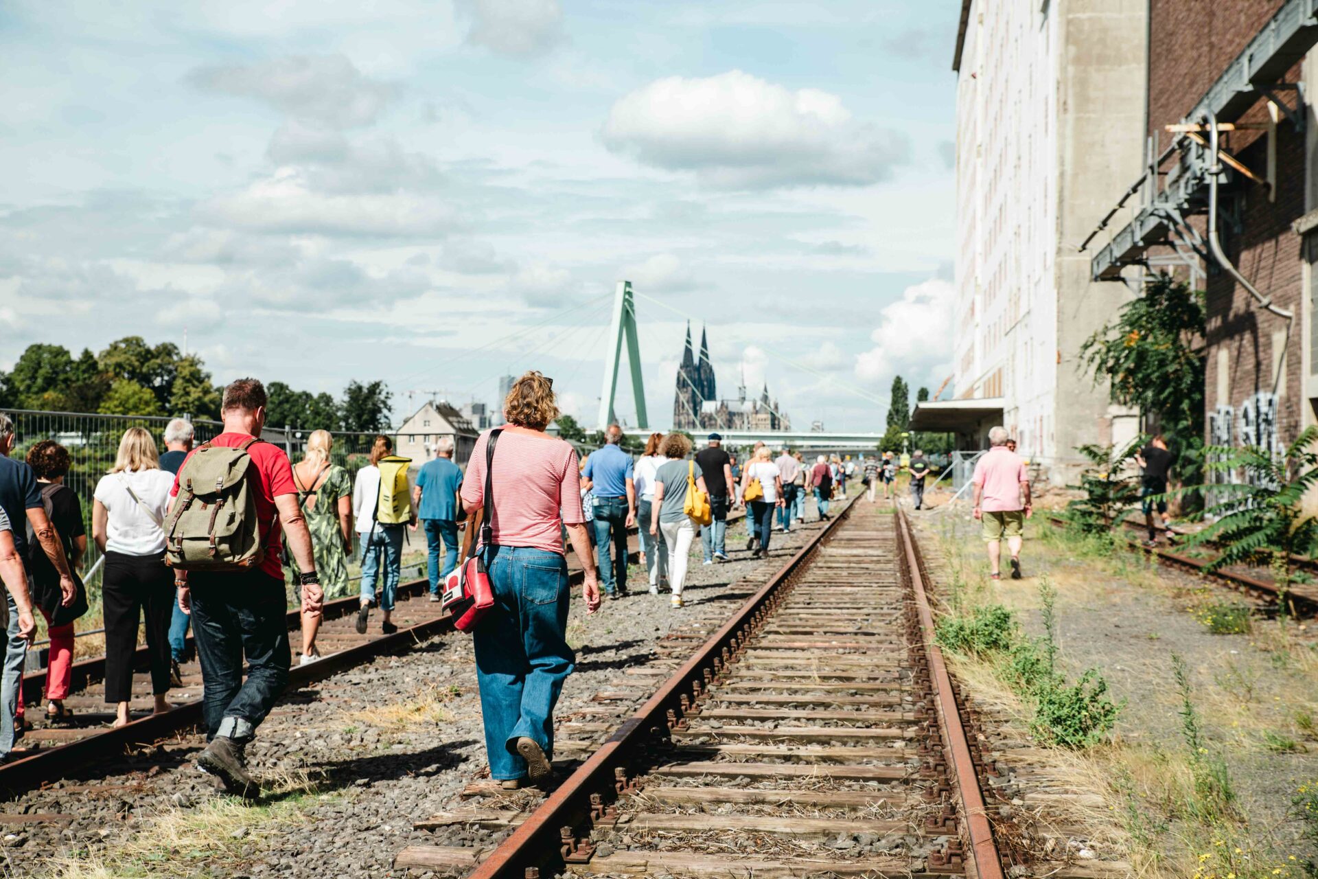 Eine große Besuchergruppe erkundet beim Hafentag 2023 das ehemalige Gleisgelände im Deutzer Hafen. Rechts verläuft ein historisches Industriegebäude, im Hintergrund ist der Kölner Dom sichtbar.